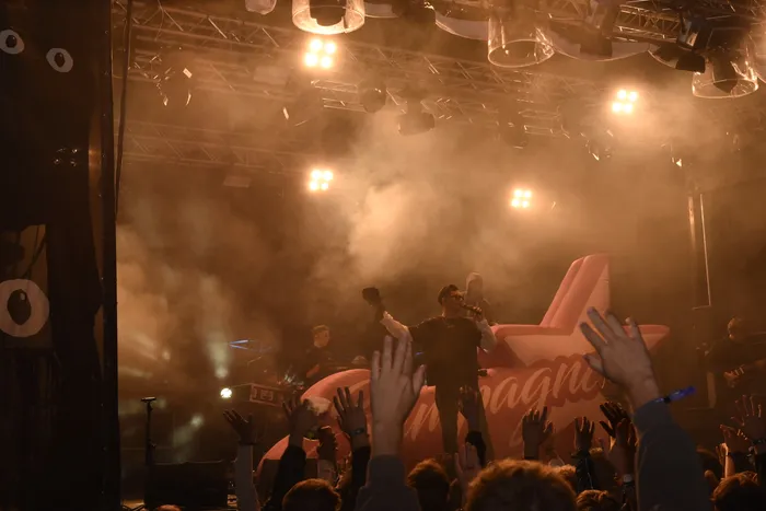 Die Band Sampagne performt auf einer vernebelten Bühne vor einem großen rosa Schriftzug, während das Publikum im Vordergrund die Hände hebt.