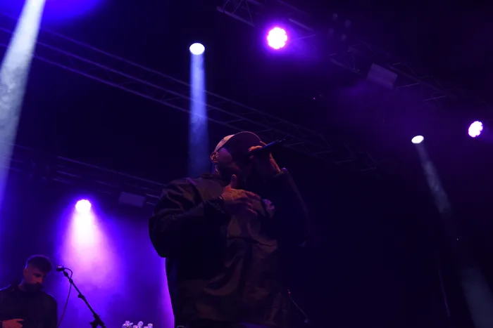 Ein Mann mit Kappe und dunkler Jacke performt mit einem Mikrofon auf einer Bühne unter violettem Scheinwerferlicht.