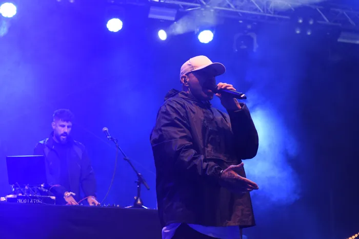 Azad rappt unter blauem Bühnenlicht in ein Mikrofon, während im Hintergrund ein DJ an einem Mischpult steht.