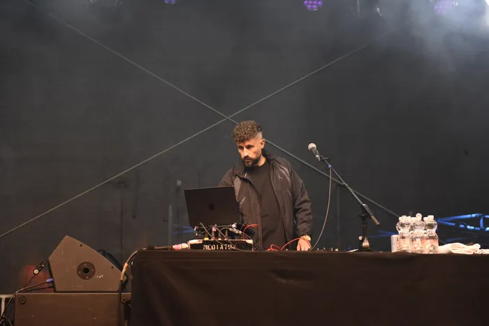 Azad bedient an einem Tisch mit Laptop und Mischpult Musik-Equipment auf einer Bühne vor dunklem, verrauchtem Hintergrund.