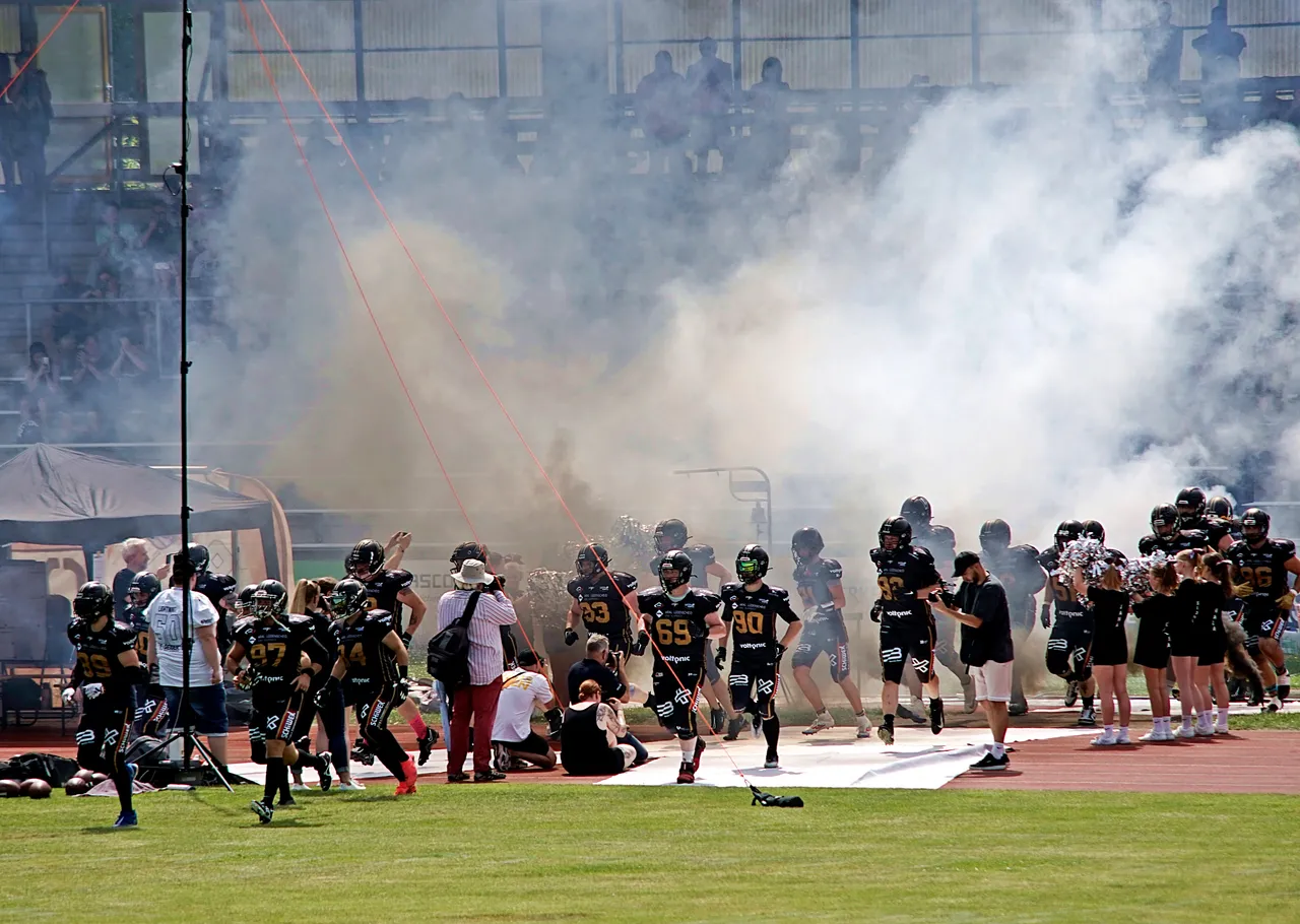 American-Football-Spieler in schwarzen Trikots betreten unter Rauchfahnen den Platz, während Cheerleader im Hintergrund stehen.
