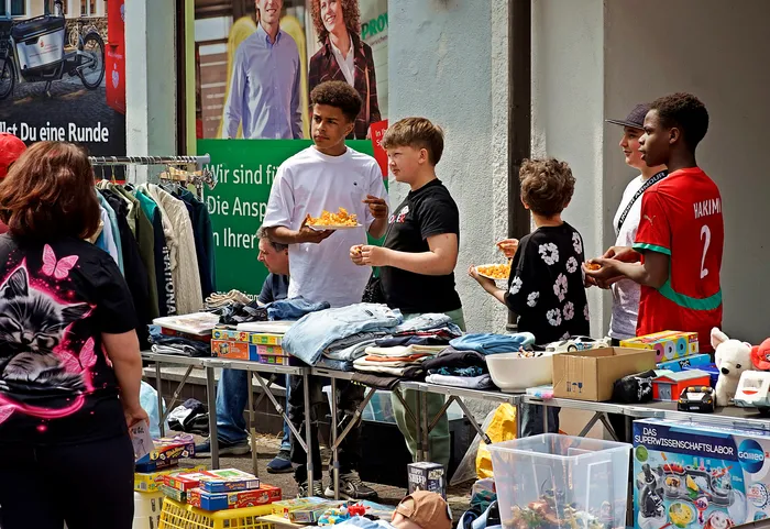 Junge Leute verkaufen auf einem Flohmarkt Kleidung, Spielzeug und andere Gegenstände. Im Hintergrund ein Plakat mit lächelnden Menschen.