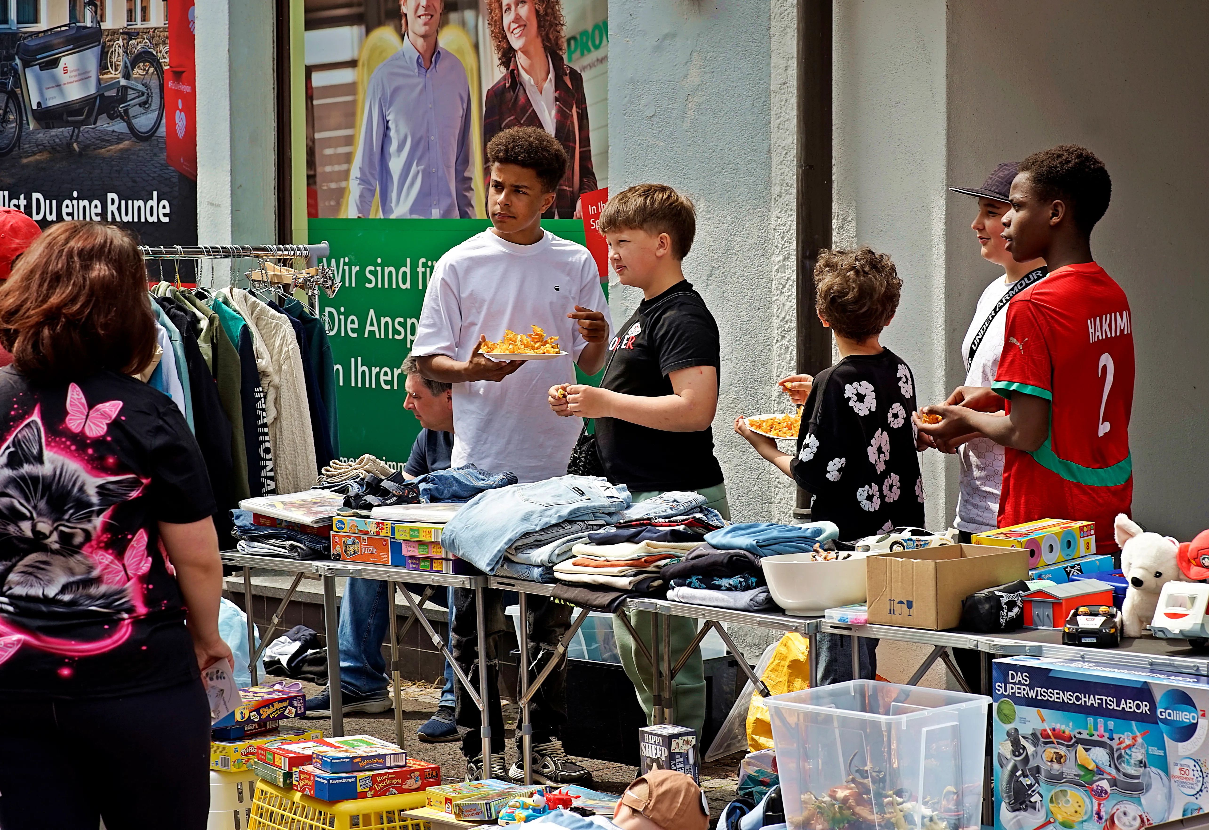 Junge Leute verkaufen auf einem Flohmarkt Kleidung, Spielzeug und andere Gegenstände. Im Hintergrund ein Plakat mit lächelnden Menschen.