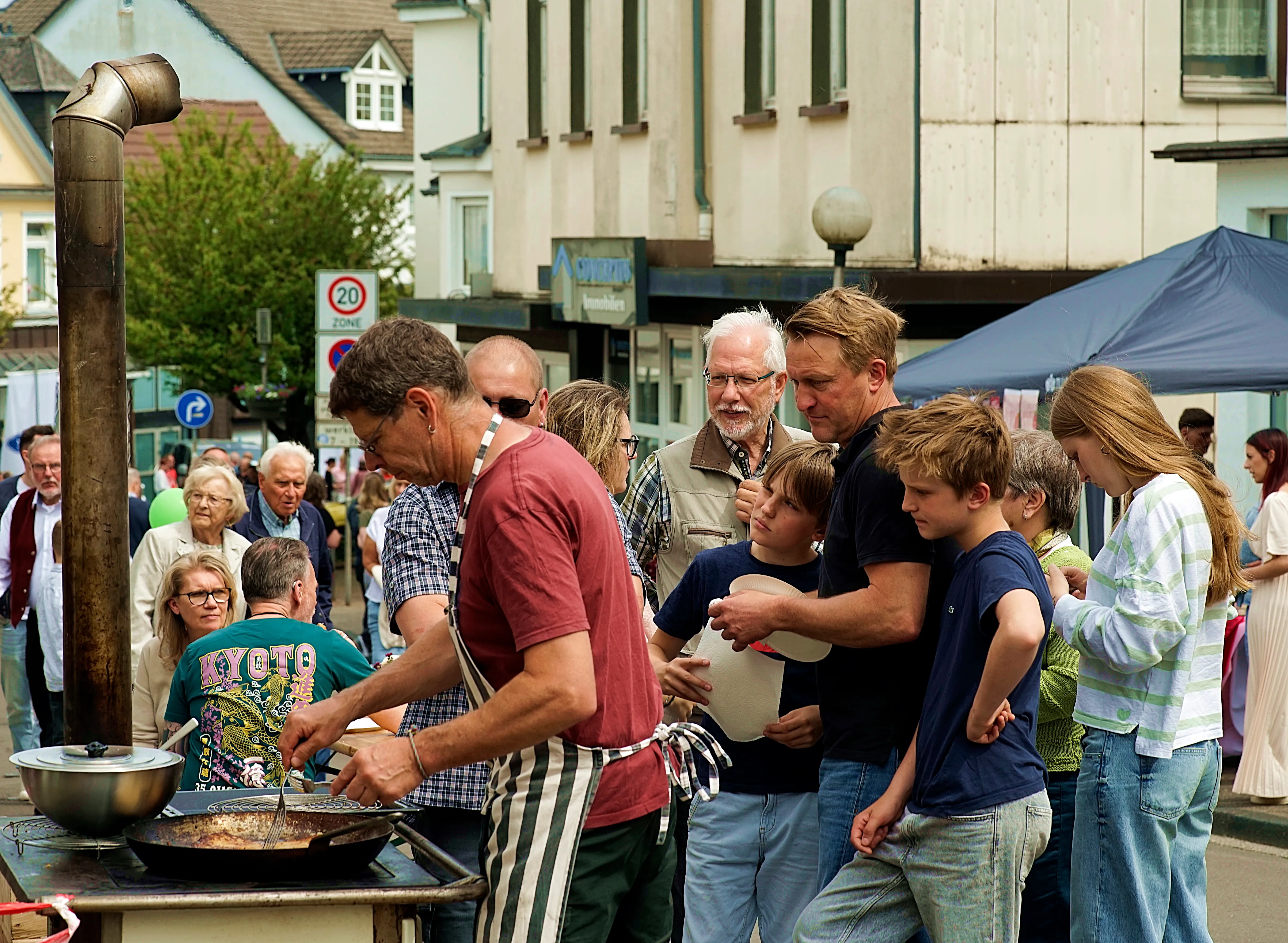 Ein Mann bereitet an einem mobilen Grill Speisen zu, während eine Gruppe von Menschen, darunter mehrere Kinder, zuschaut und sich in einer Fußgängerzone aufhält.