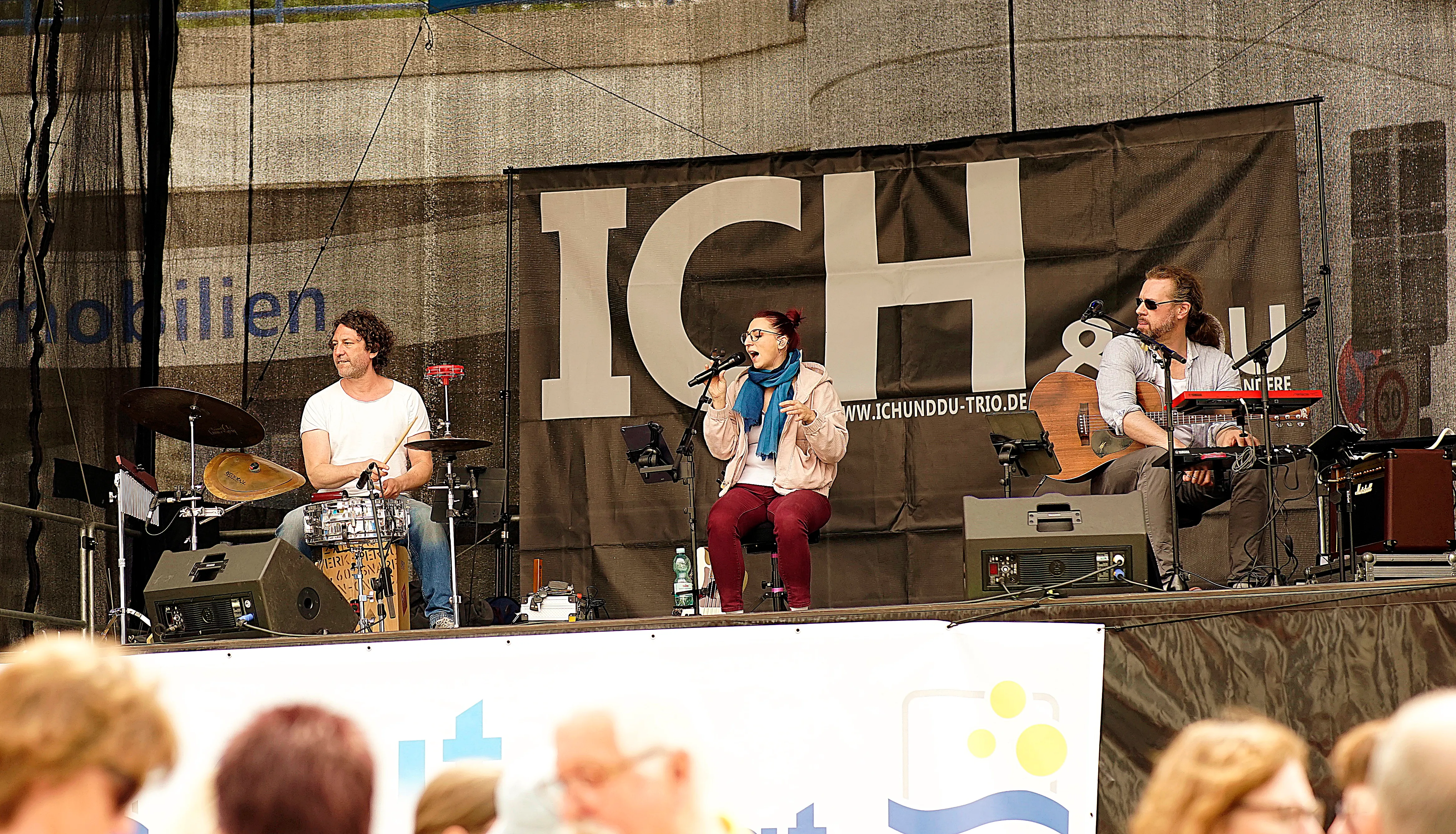 Drei Musiker spielen auf einer Bühne vor Publikum. Im Hintergrund ein Banner mit der Aufschrift „ICH & DU“ und einer Website-Adresse.