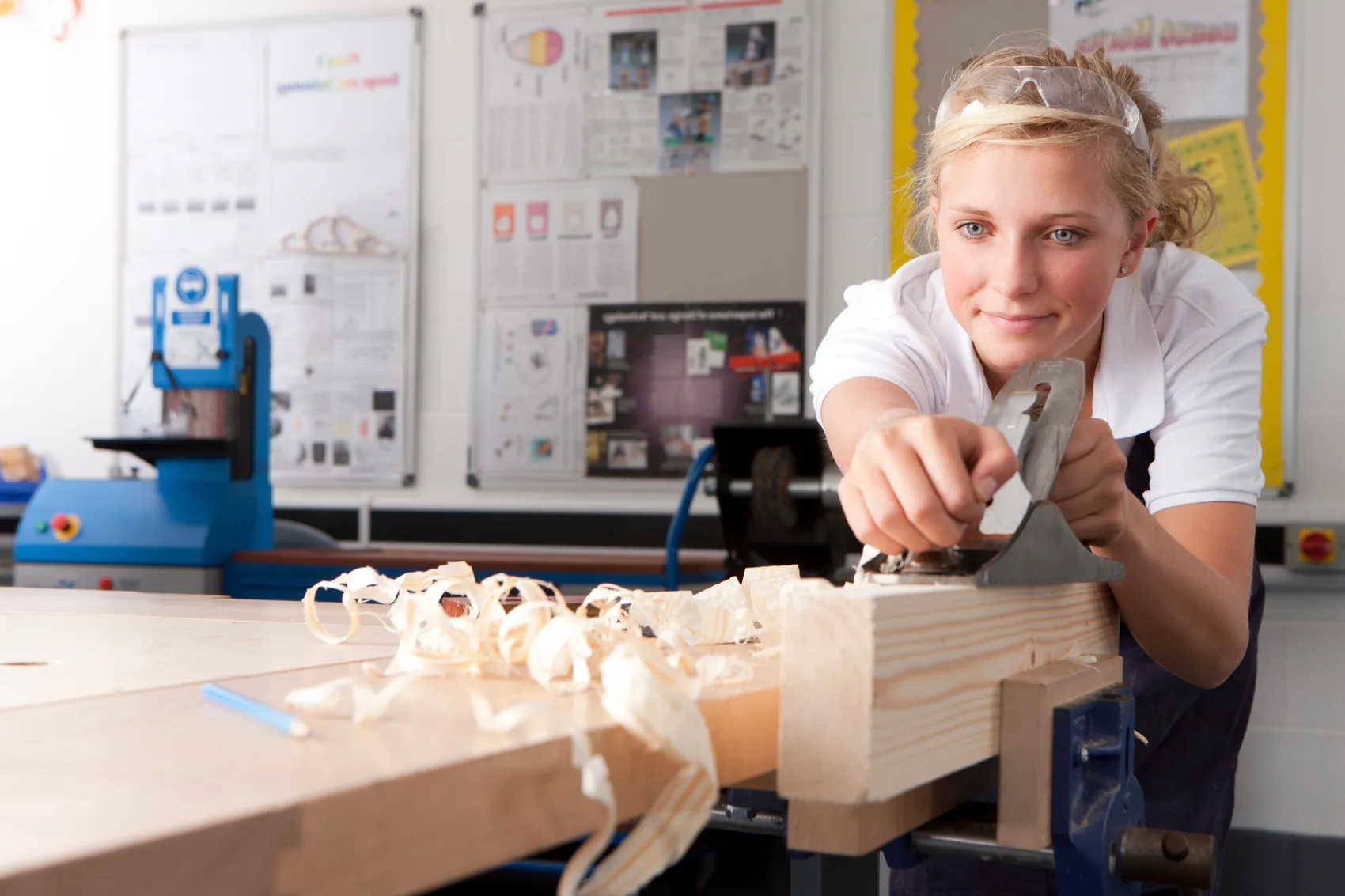 Junge Frau bearbeitet mit einem Hobel ein Holzstück in einer Werkstatt. Im Hintergrund sind Werkzeuge und Plakate zu sehen.