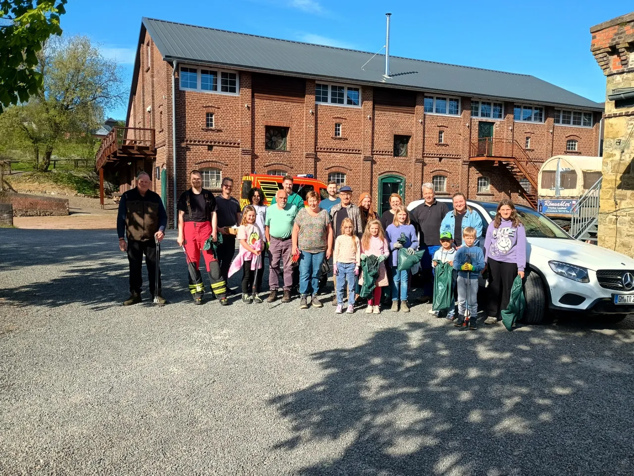 Eine Gruppe von etwa zwanzig Personen steht vor einem zweistöckigen Backsteingebäude mit grauem Dach. Im Hintergrund steht ein silberner PKW und ein rotes Feuerwehrauto.