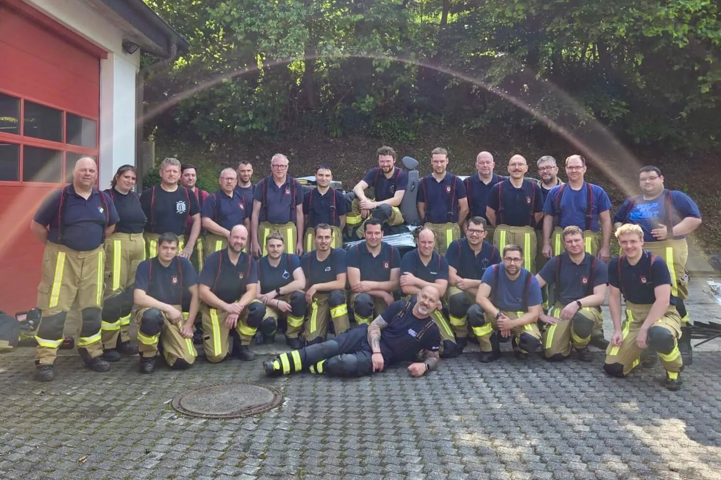 Gruppe von Feuerwehrleuten in Uniformen posiert vor einem Feuerwehrhaus, während ein Wasserstrahl im Hintergrund zu sehen ist.