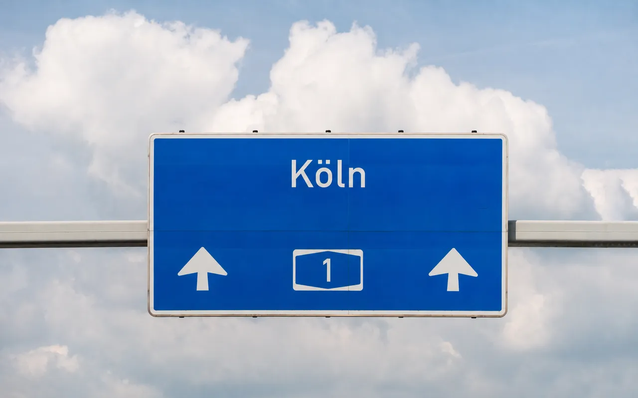 Blaues Autobahnschild mit der Aufschrift „Köln“, zwei weißen Pfeilen nach oben und der Ziffer 1 vor einem bewölkten Himmel.