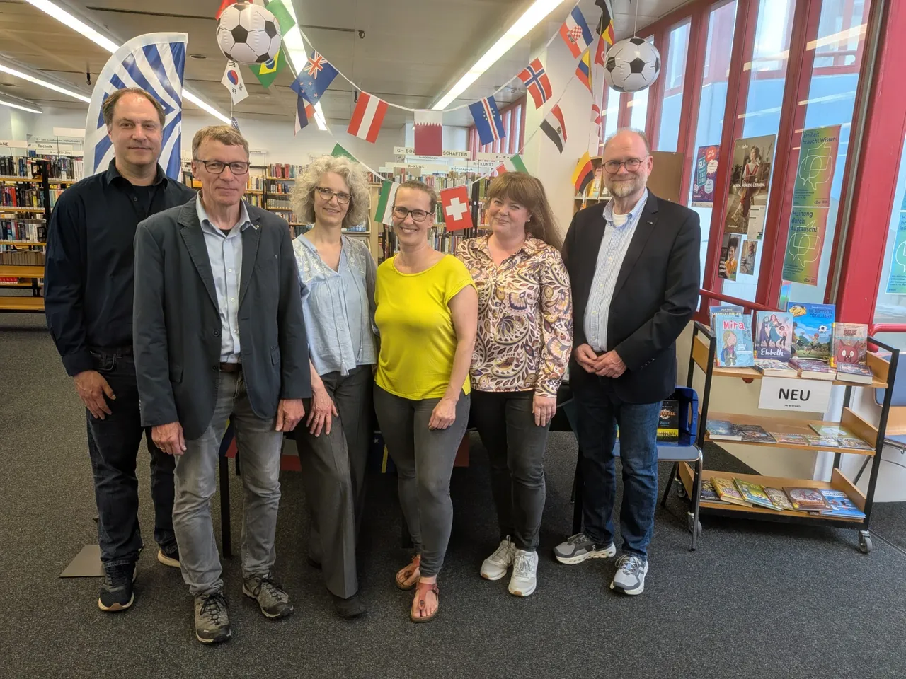 Sechs Personen stehen in einer Bibliothek vor Regalen mit Büchern und einem Rollwagen mit neuen Büchern. Die Gruppe besteht aus drei Männern und drei Frauen, die alle lächeln und in formeller Kleidung gekleidet sind. Im Hintergrund sind Flaggen und ein Fußball zu sehen.