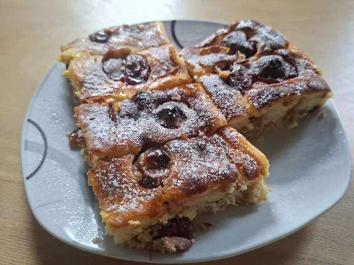 Mehrere Stücke Pflaumenkuchen mit dunklen Pflaumen und Puderzucker auf einem weißen Teller.