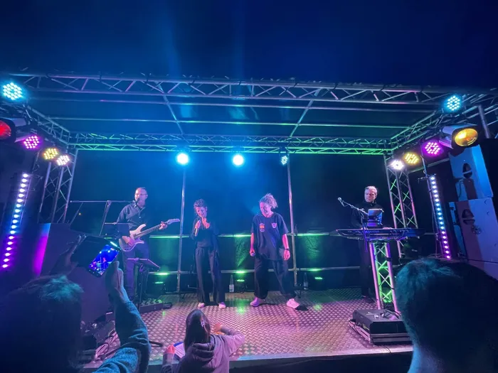 Eine Band spielt auf einer Bühne vor Publikum. Ein Sänger mit Mikrofon steht im Zentrum, flankiert von Musikern mit Gitarre und Keyboard unter blauem und grünem Licht.