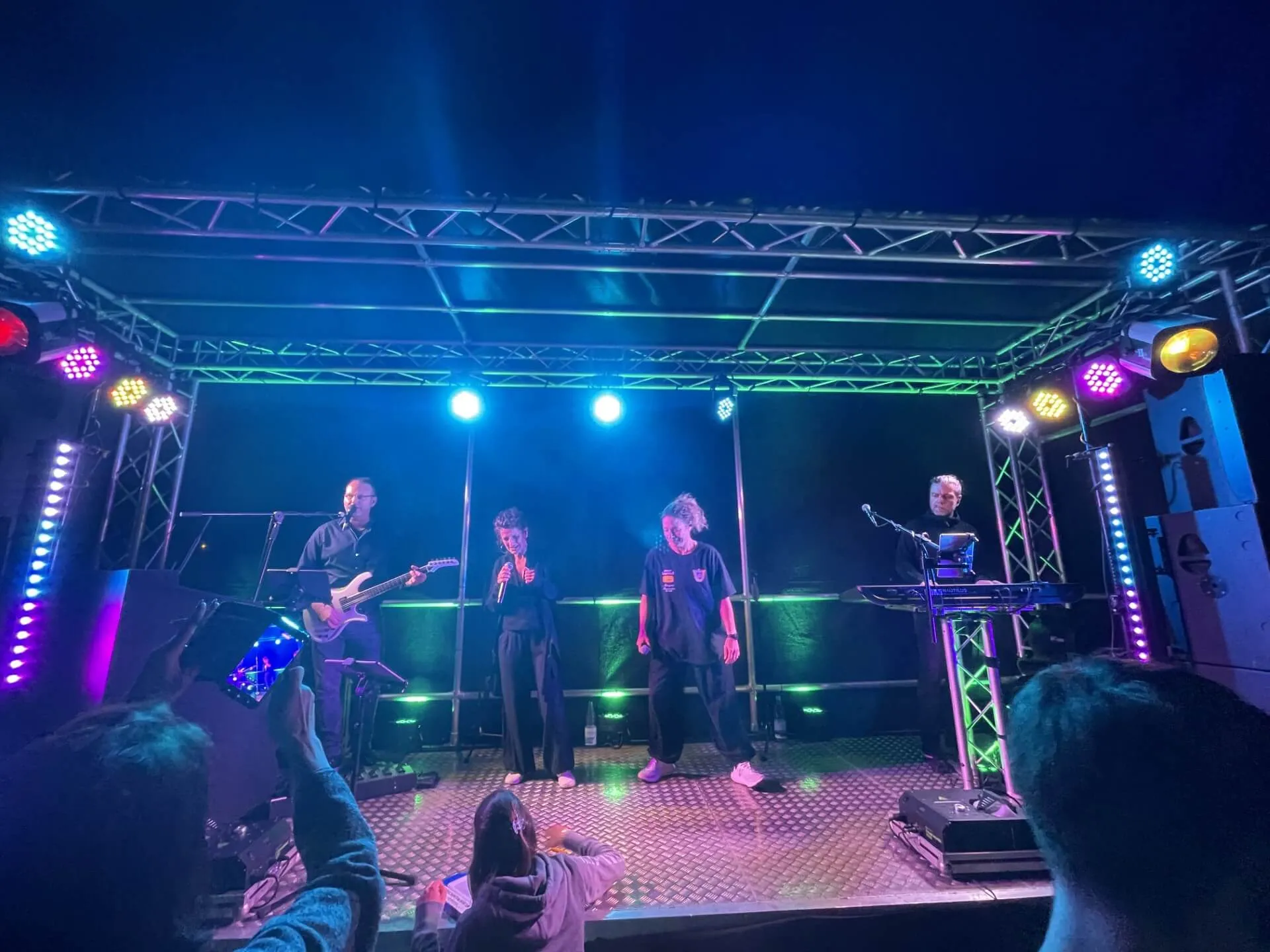 Eine Band spielt auf einer Bühne vor Publikum. Ein Sänger mit Mikrofon steht im Zentrum, flankiert von Musikern mit Gitarre und Keyboard unter blauem und grünem Licht.