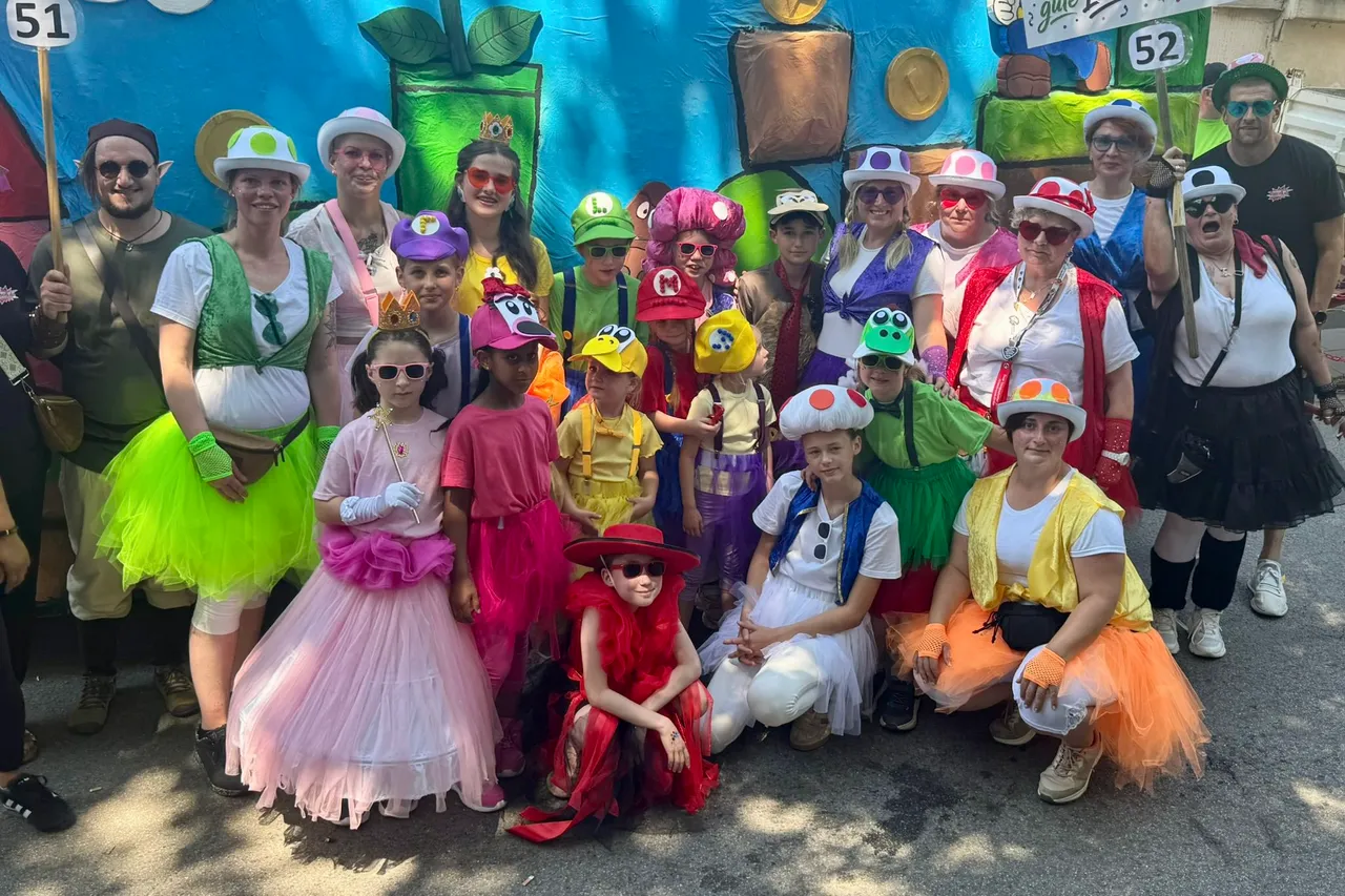 Eine große Gruppe von Kindern und Erwachsenen in farbenfrohen Kostümen, inspiriert von Figuren aus dem Super Mario Universum, posiert für ein Foto während eines Umzugs. Viele tragen Hüte und Accessoires, die an die Charaktere erinnern. Im Hintergrund sind weitere Teilnehmer und Dekorationen zu sehen.