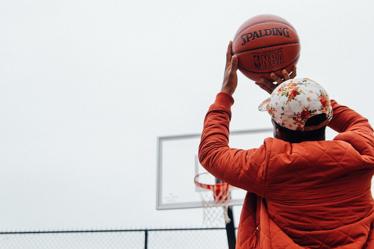 Ein Basketballspieler wirft den Ball auf einen Korb. Er trägt eine rote Jacke und eine blumige Kappe. Im Hintergrund ist ein Basketballfeld zu sehen.