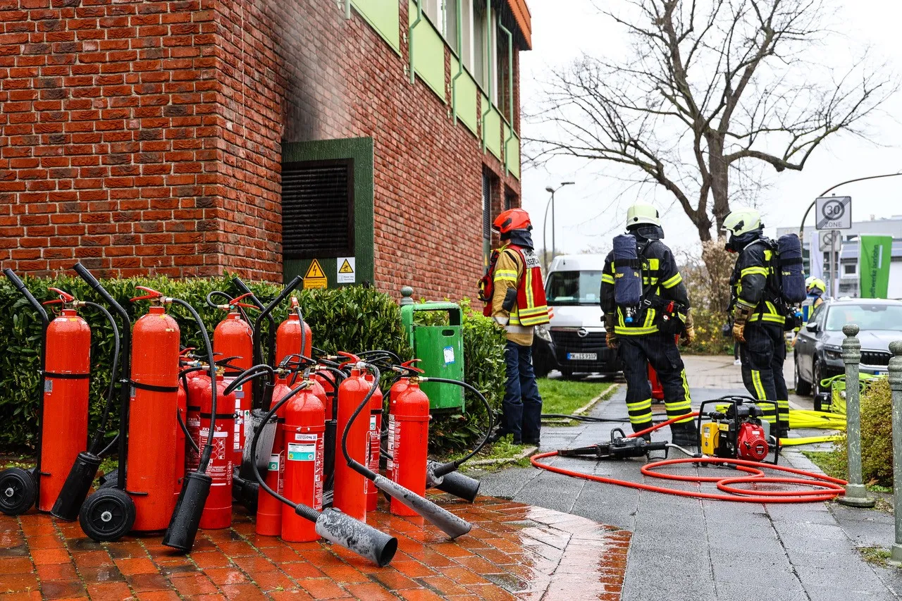 Mehrere rote CO2-Feuerlöscher stehen vor einer Backsteinfassade, während Feuerwehrleute im Hintergrund arbeiten und einen Löschschlauch vorbereiten.