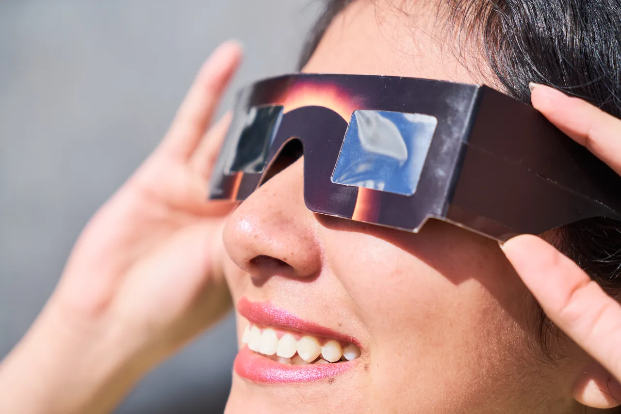 Eine lächelnde Frau trägt eine dunkle Sonnenfinsternis-Brille und hält diese mit den Händen fest.
