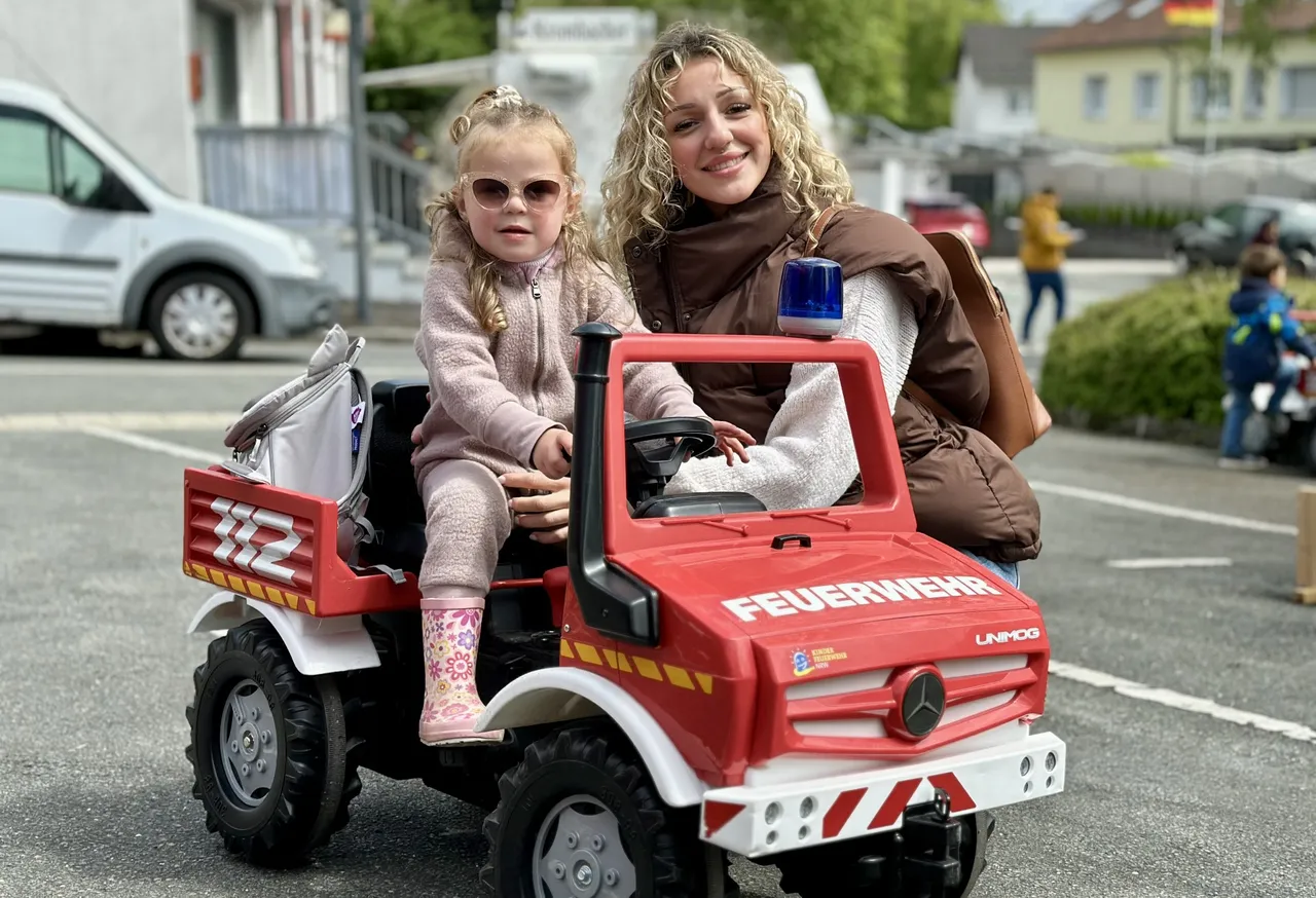 Ein kleines Mädchen mit Sonnenbrille sitzt auf einem roten Feuerwehrauto, während eine Frau neben ihr lächelt. Im Hintergrund sind weitere Menschen und Gebäude zu sehen.