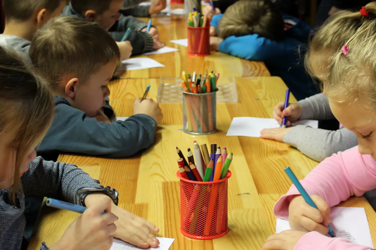 Kinder sitzen an einem Holztisch und malen oder schreiben. Zwischen ihnen stehen Becher mit Stiften und Buntstiften.