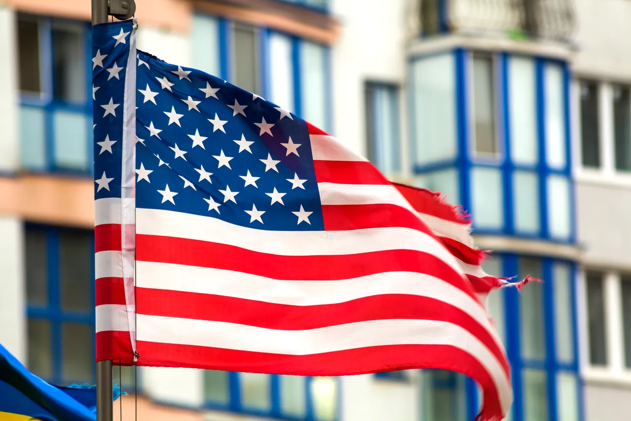 Die amerikanische Flagge weht vor einem modernen Gebäude mit blauen Fenstern. Die Flagge zeigt rote und weiße Streifen sowie einen blauen Bereich mit weißen Sternen.