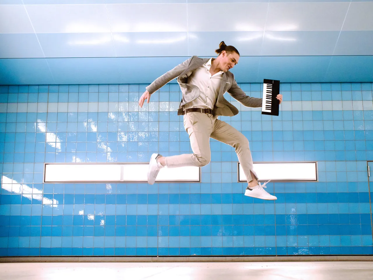 Kabarettist Andreas Langsch springt mit einem kleinen Keyboard in der Hand vor einer blau gefliesten Wand.
