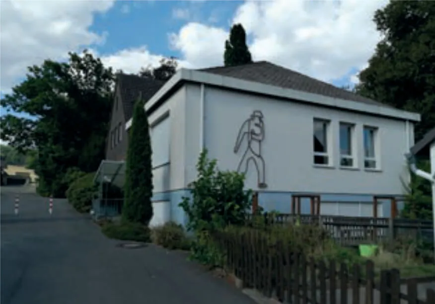 Helles zweigeschossiges Gebäude mit Flachdach und großem Wandgemälde eines Mannes mit Hut und Koffer. Vor dem Gebäude verläuft eine Straße und ein Holzzaun.