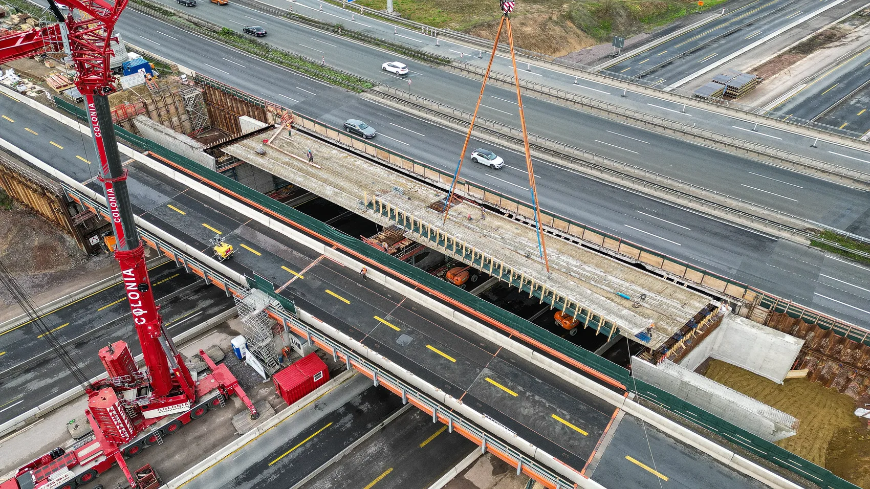 Eine neue Brücke wird über eine Autobahn gehoben. Zwei große Kräne sind im Einsatz, um das Brückenteil zu positionieren.