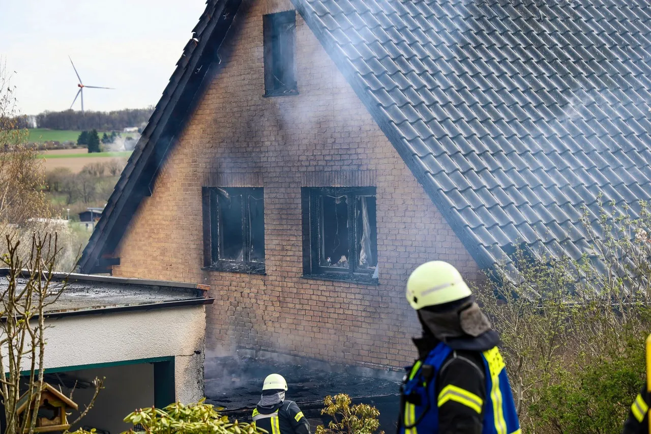 Ein Wohnhaus mit ausgebrannten Fenstern und Rauch, nachdem ein Feuer von einem Carport auf das Gebäude übergegriffen hat. Feuerwehrleute stehen im Vordergrund.