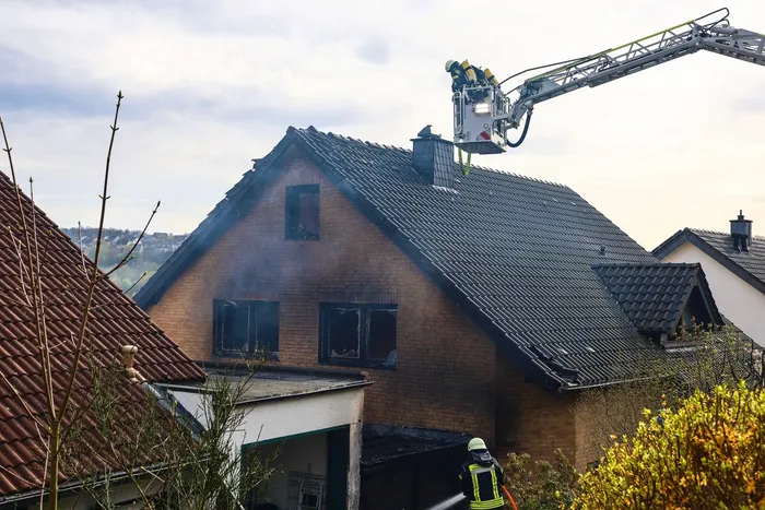 Feuerwehrleute bekämpfen einen Brand in einem zweigeschossigen Wohnhaus mit dunklem Dach. Rauch quillt aus den Fenstern des Hauses.