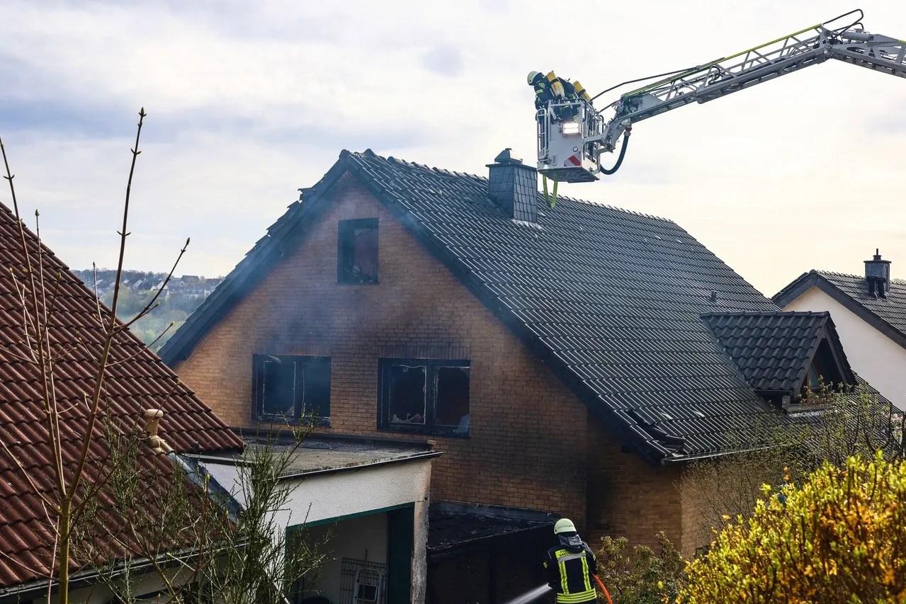 Feuerwehrleute bekämpfen einen Brand in einem zweigeschossigen Wohnhaus mit dunklem Dach. Rauch quillt aus den Fenstern des Hauses.