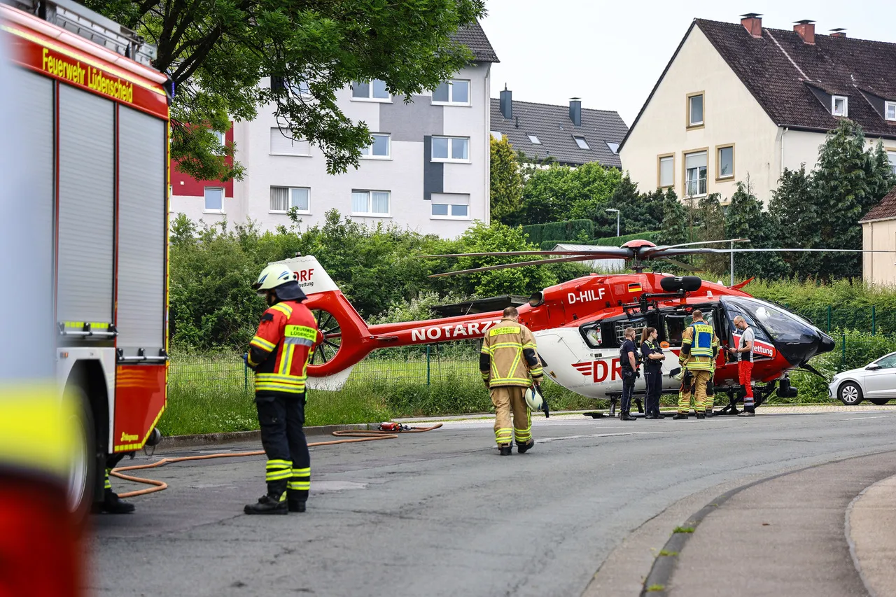 Rettungskräfte stehen um einen roten Rettungshubschrauber mit der Aufschrift „Notarzt“ und „D-HILF“. Im Hintergrund sind Wohngebäude und ein rotes Feuerwehrauto zu sehen.
