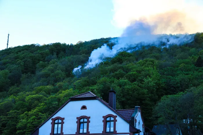 Rauch steigt über dichtem Wald auf, im Vordergrund ein rotdachiges Gebäude.