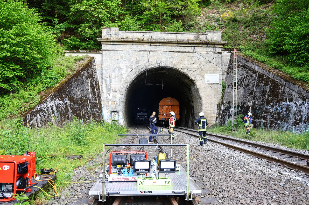 Zug fährt aus einem Tunnel, während mehrere Einsatzkräfte in gelben Westen am Gleis stehen. Im Vordergrund ein rotes Wartungsfahrzeug mit Scheinwerfern.