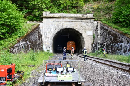 Zug fährt aus einem Tunnel, während mehrere Einsatzkräfte in gelben Westen am Gleis stehen. Im Vordergrund ein rotes Wartungsfahrzeug mit Scheinwerfern.