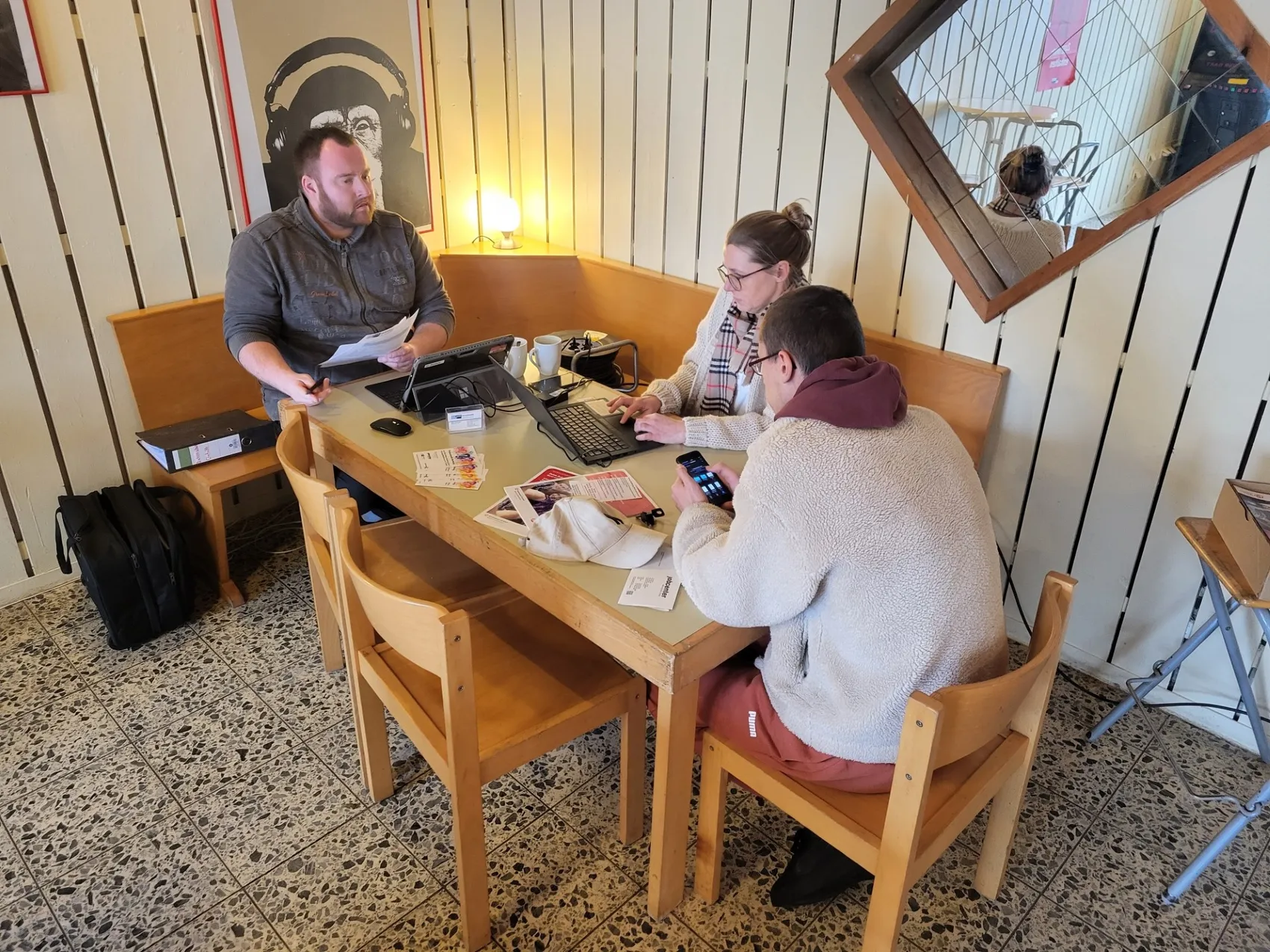 Drei Personen sitzen an einem Holztisch in einem Raum mit roten Wänden und diskutieren. Eine Person tippt auf einem Laptop, während die anderen beiden Smartphones betrachten. Im Hintergrund befindet sich ein Spiegel und eine kleine Lampe.