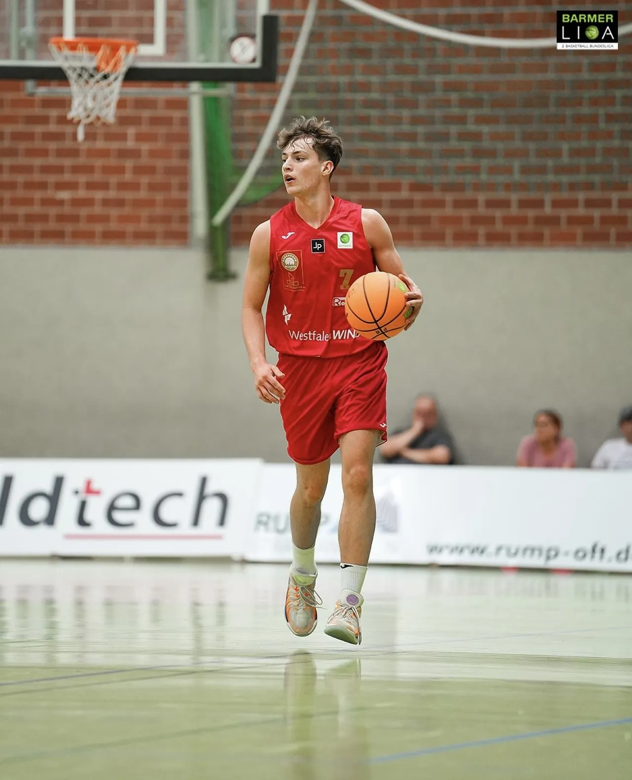 Ein Basketballspieler in roter Trikot und Shorts dribbelt den Ball auf einem Hallenparkett. Im Hintergrund sind Zuschauer und Werbebanner zu sehen.