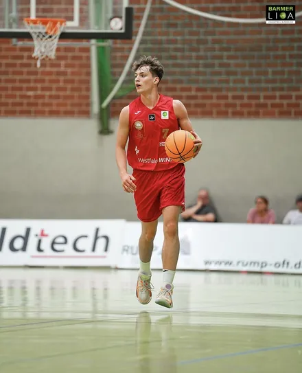 Ein Basketballspieler in roter Trikot und Shorts dribbelt den Ball auf einem Hallenparkett. Im Hintergrund sind Zuschauer und Werbebanner zu sehen.