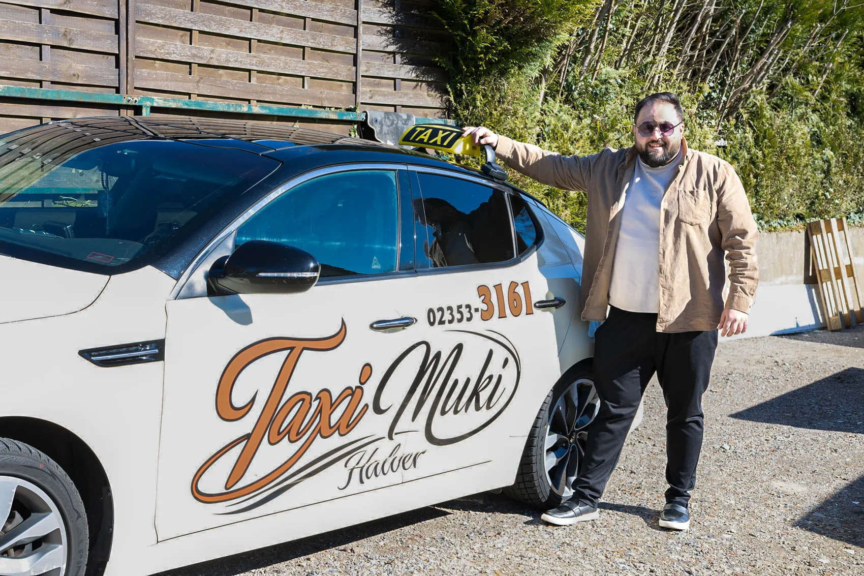 Muhsin Aksu, Inhaber des Taxiunternehmens „Taxi Muki“ in Halver, steht lächelnd neben einem Taxi mit dem Schriftzug und der Telefonnummer des Unternehmens.
