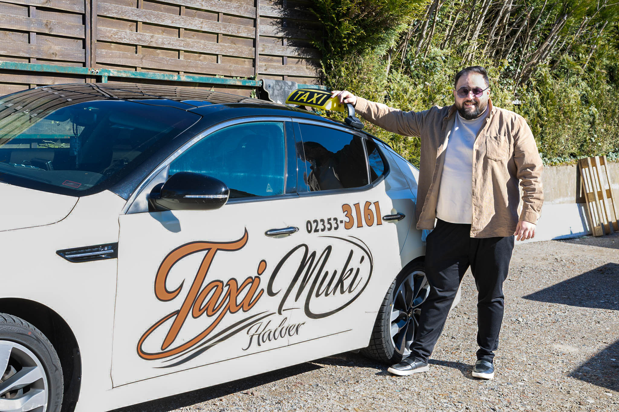 Muhsin Aksu, genannt „Muki“, steht lächelnd neben seinem Taxi mit der Aufschrift „Taxi Muki Halver“ und deutet auf das Taxischild.