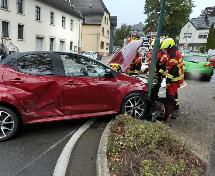 Ein rotes Auto mit massiven Unfallschäden an der Seite und Front, während Feuerwehrleute am Fahrzeug arbeiten.
