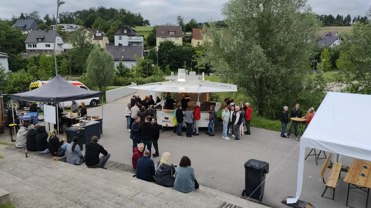 Eine kleine Menschenmenge steht und sitzt vor einem Imbisswagen und einem Pavillon in einem Park. Im Hintergrund sind Wohnhäuser und Bäume zu sehen.