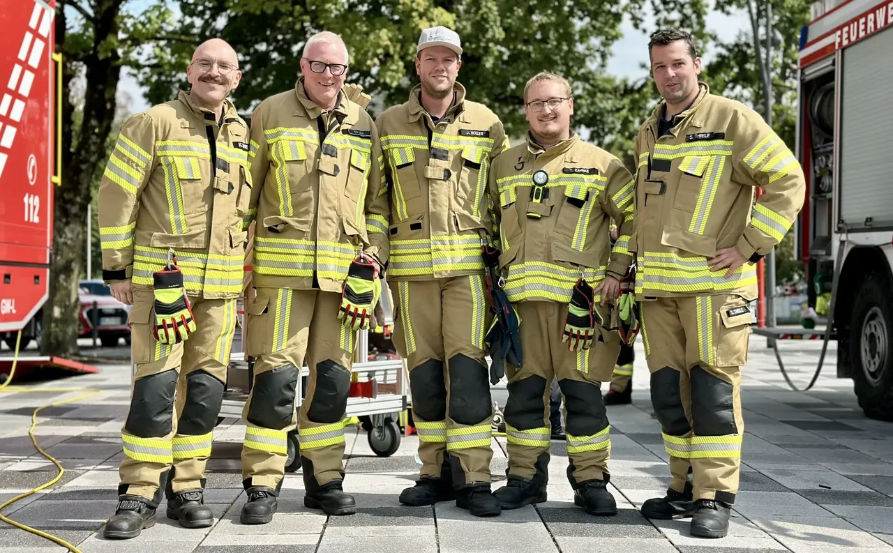 Fünf Feuerwehrleute in gelb-beiger Schutzkleidung mit Reflexstreifen stehen nebeneinander vor einem Feuerwehrfahrzeug.