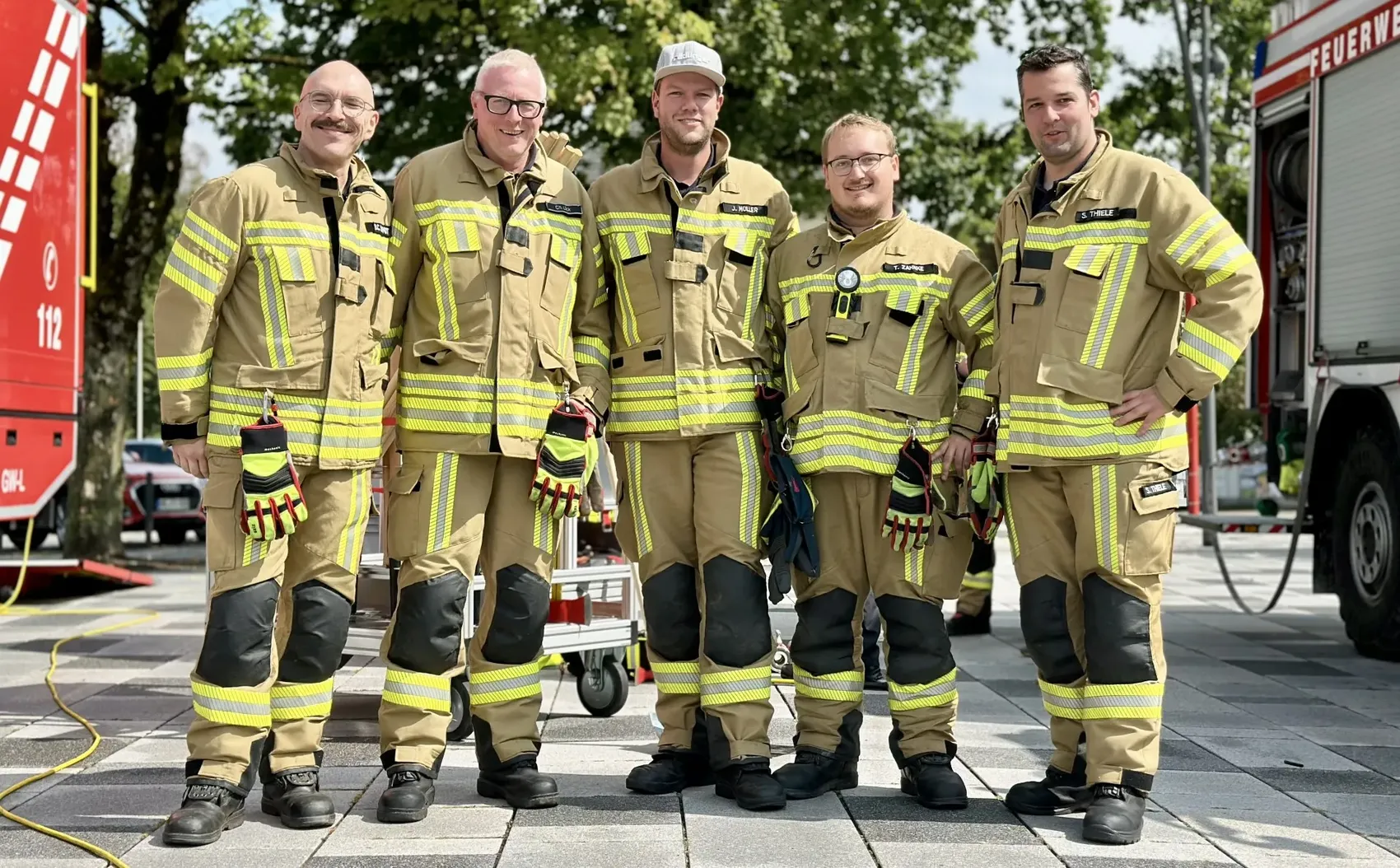 Fünf Feuerwehrleute in gelb-beiger Schutzkleidung mit Reflexstreifen stehen nebeneinander vor einem Feuerwehrfahrzeug.