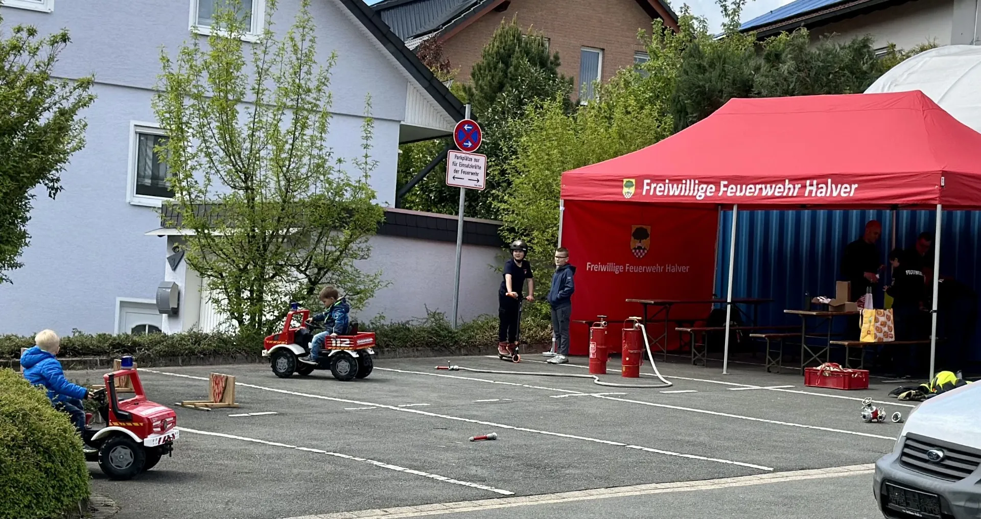 Kinder fahren mit Spielzeug-Feuerwehrautos vor einem Stand der Freiwilligen Feuerwehr Halver, während ein Feuerwehrmann im Hintergrund steht.