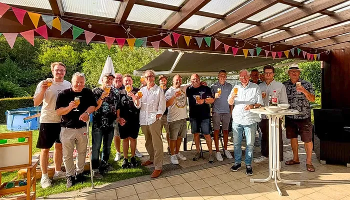 Eine Gruppe von Männern steht unter einer Holzpergola mit bunten Wimpelketten und hält Gläser mit Bier hoch.