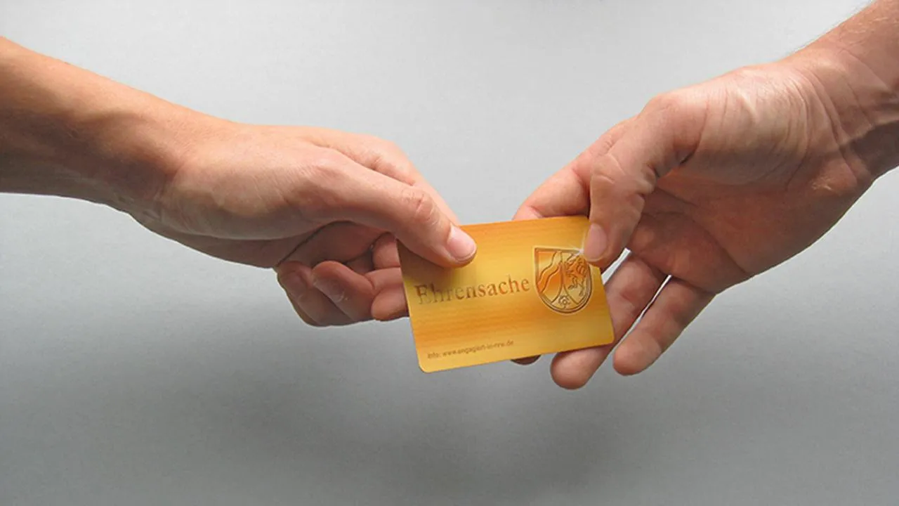 Zwei Hände übergeben sich eine gelbe Ehrenamtskarte mit dem Schriftzug „Ehrensache“ und einem Wappen.