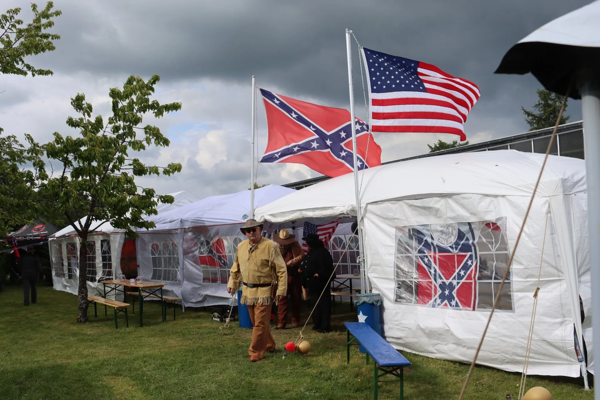 Zwei Männer in Western-Kleidung gehen an Ständen vorbei, die mit der Flagge der Konföderierten Staaten und der US-Flagge geschmückt sind. Im Hintergrund sind weitere Zelte und ein bewölkter Himmel zu sehen.