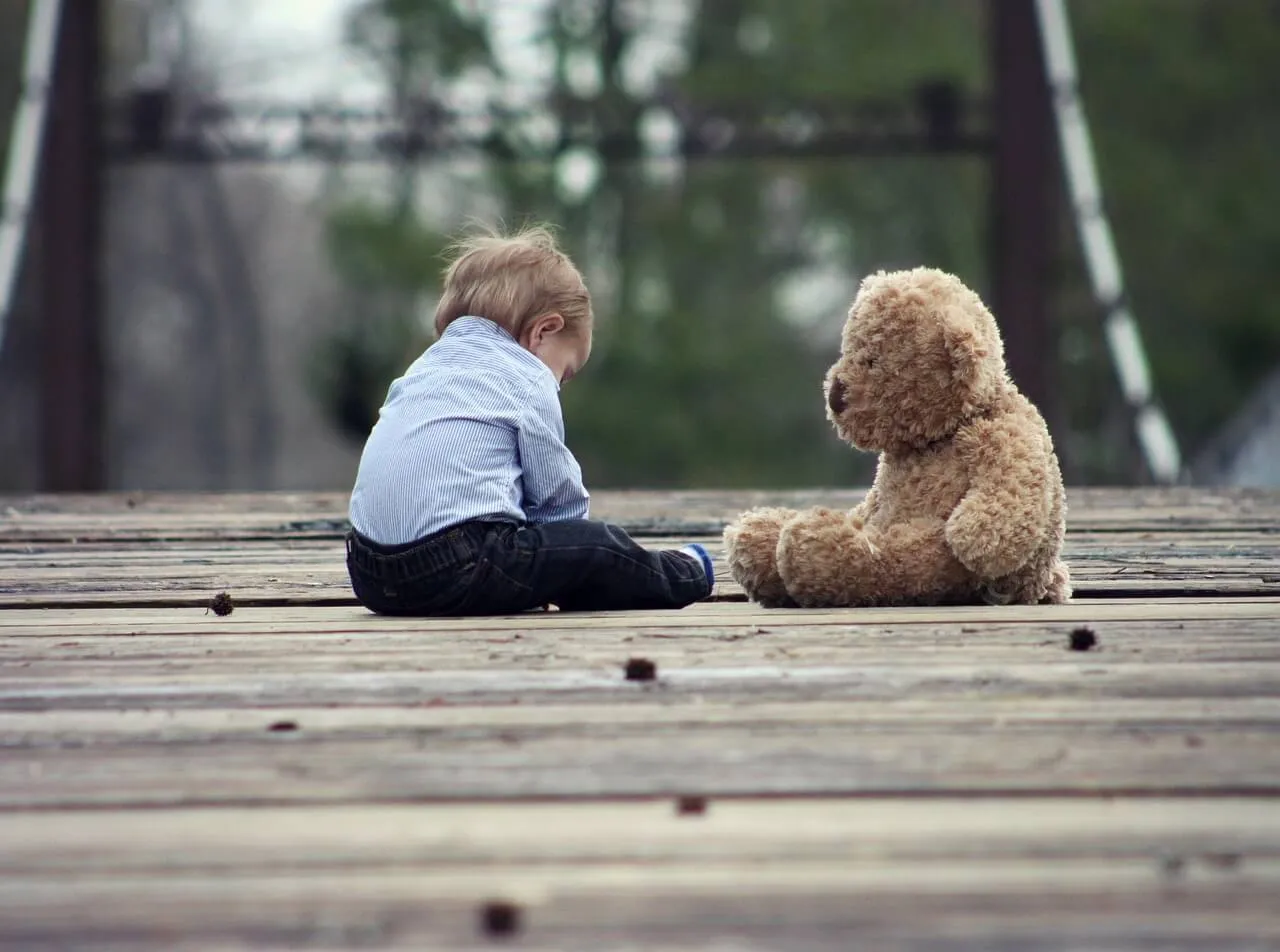 Ein Kind in einem hellblau gestreiften Hemd sitzt auf einem Holzsteg und blickt einem großen, hellbraunen Teddybären gegenüber.