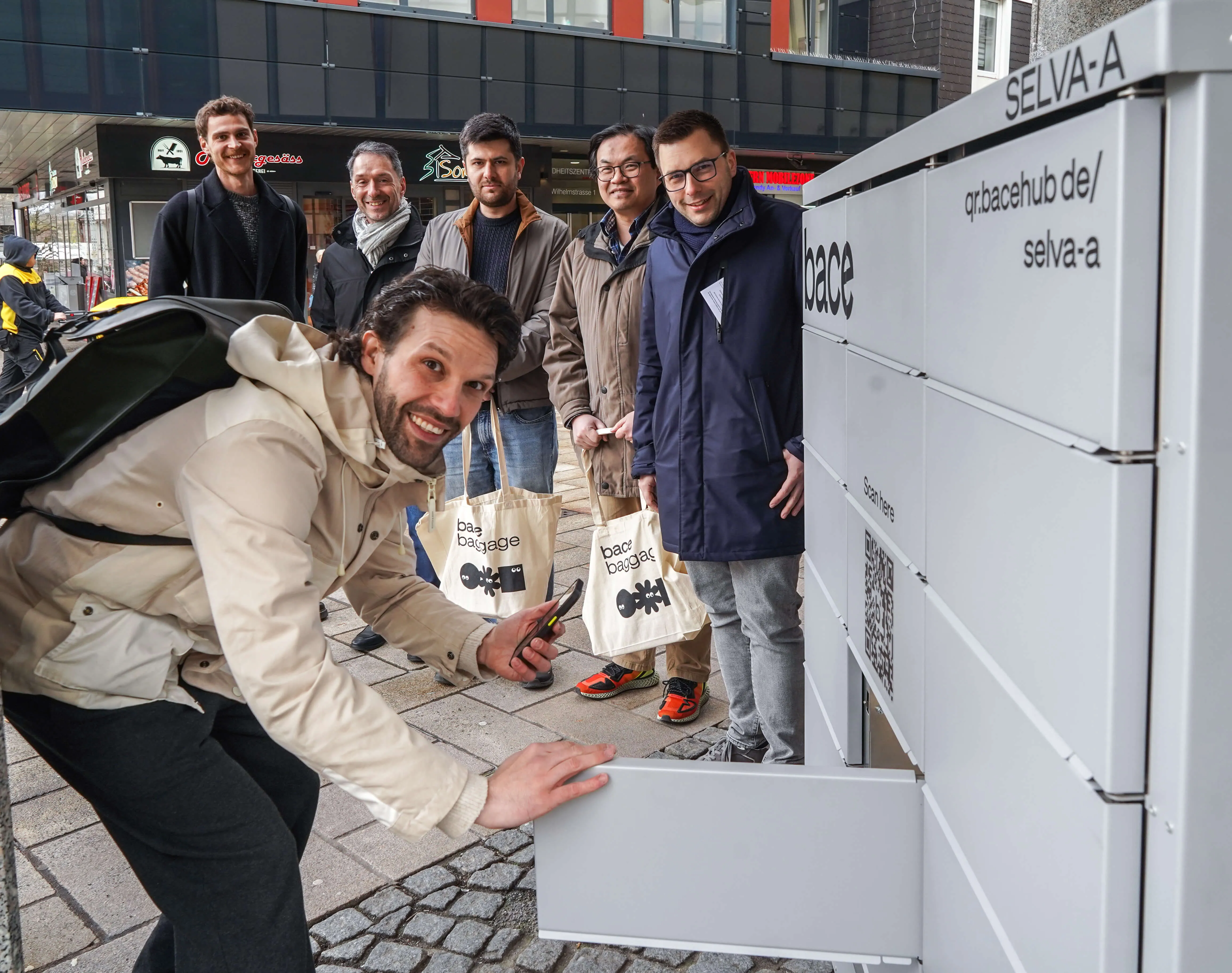 Ein bace-Mitarbeiter demonstriert die Nutzung eines Gepäckschließfachs, während weitere Personen im Hintergrund zusehen. Im Hintergrund sind Geschäfte und ein moderner Gebäudekomplex zu erkennen.