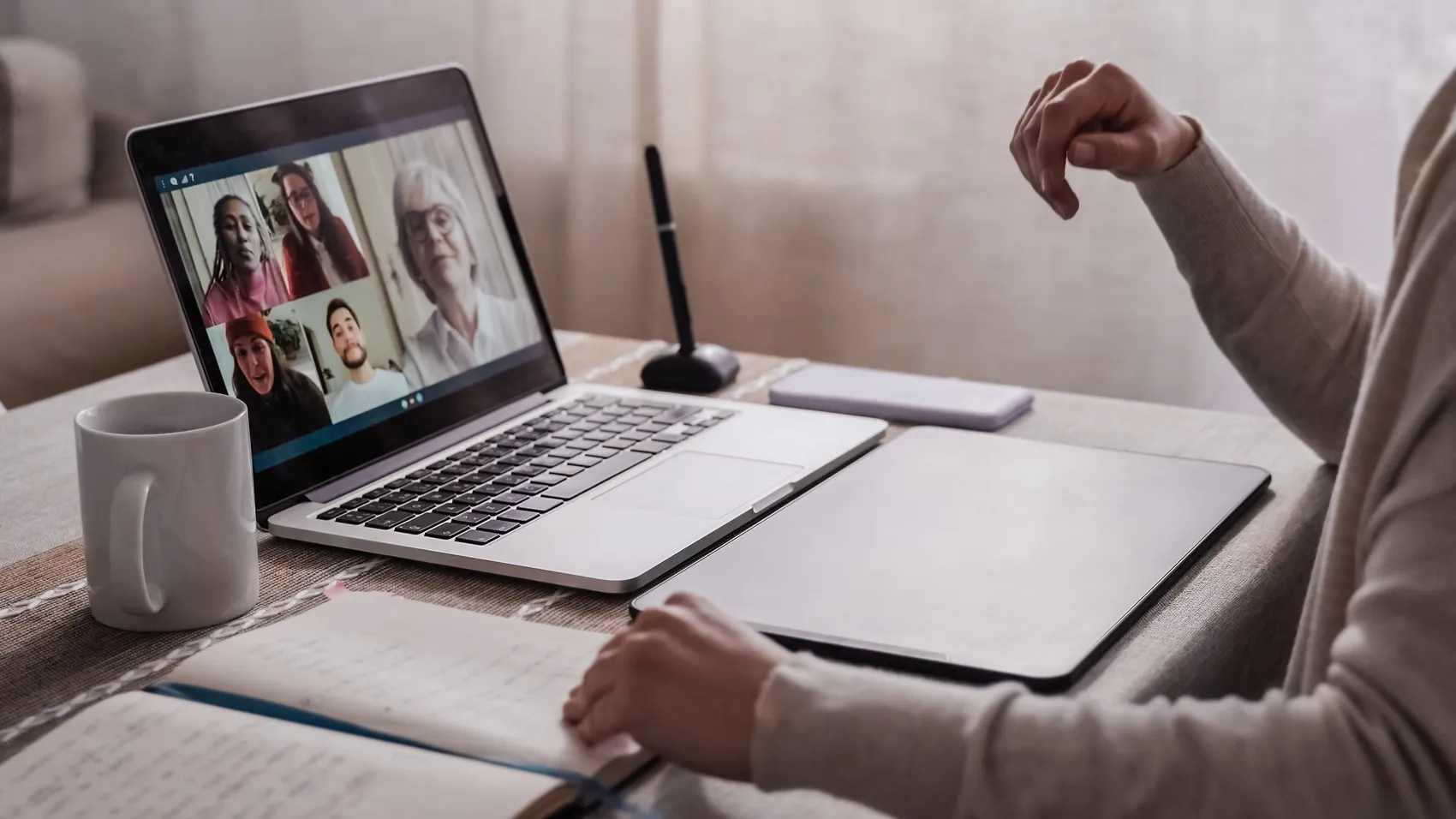 Eine Person sitzt mit dem Rücken zum Betrachter vor einem geöffneten Laptop. Auf dem Bildschirm sind vier Personen in einer Videokonferenz zu sehen.